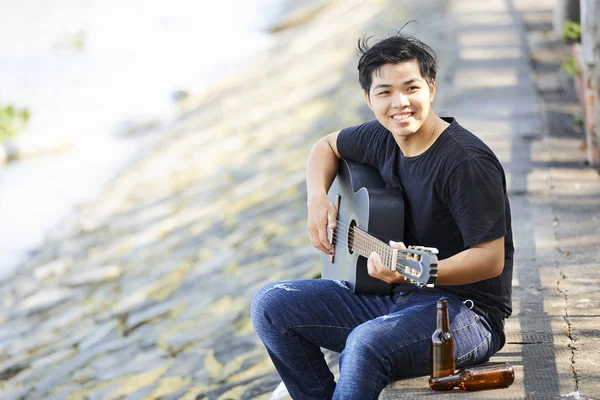 アジアの若者がギターを弾き 微笑むビールの空のボトルで屋外で休んでいる — ストック写真