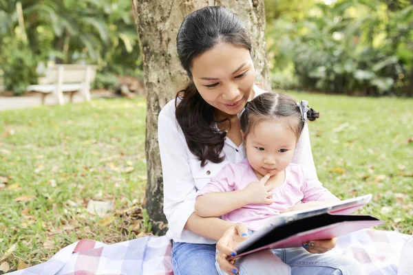 亚洲母亲和女儿坐在城市公园的树下 在数字平板电脑上观看卡通片 — 图库照片