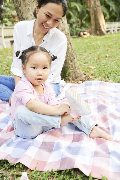 彼女は都市公園で毛布の上に座っているように 小さな娘がスニーカーを脱ぐのを見て幸せな母親 — ストック写真