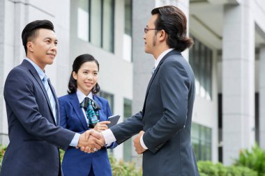 Takım elbiseli iki Asyalı işadamı ayakta ve el sıkışarak kadın asistan onlarla açık havada duran anlaşma kabul etmek
