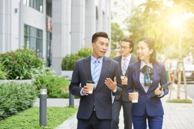 Bir grup iş adamı tek kullanımlık bardaklardan kahve içip sokakta birlikte yürürken konuşuyorlar.