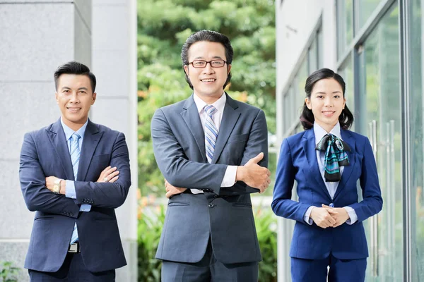 Portrait of Asian smiling businessman in eyeglasses standing in suit with arms crossed together with his business team outdoors