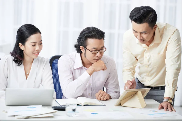亚洲年轻男性领袖在商务会议桌旁向业务合作伙伴展示数字平板电脑上的在线演示 — 图库照片