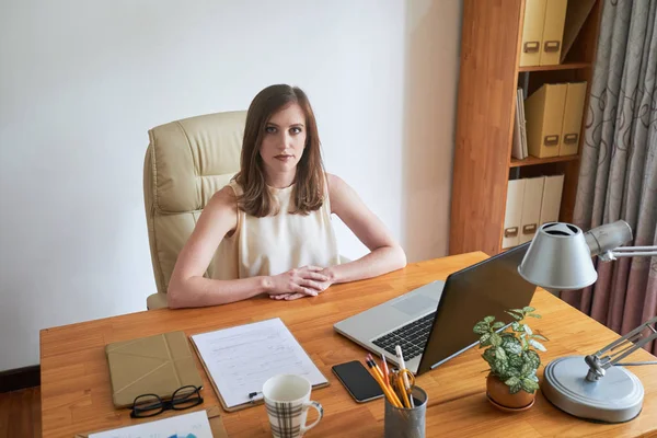 Portrait of young serious manager sitting at her workplace with laptop and documents and looking at camera at office