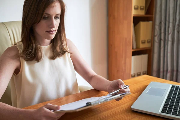 Serious businesswoman sitting at the table in front of laptop with clipboard in her hands and reading a business document