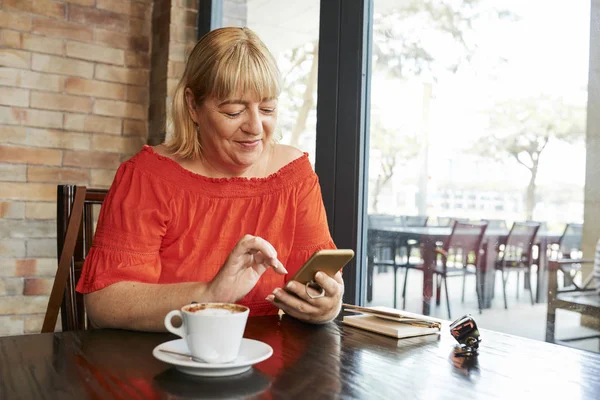 Lächelnde Reife Frau Sitzt Cafétisch Und Liest Nachrichten Ihrem Telefon — Stockfoto
