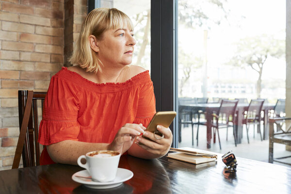 Sad pensive woman with smartphone in hands sitting at cafe table and looking through window