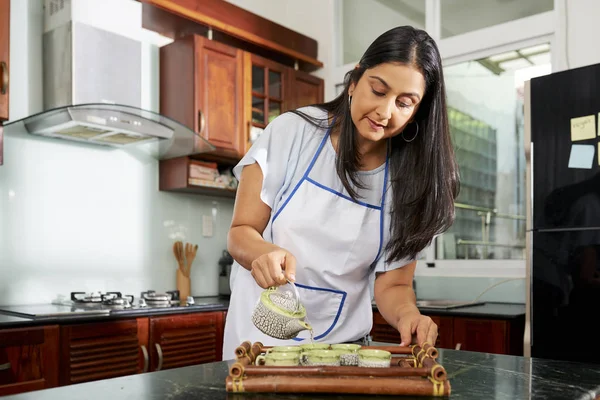 Woman in white apron pouring tea in small ceramic cups on wooden tray