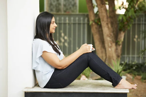 Pensive young Indian woman sitting on porch and enjoying calm morning with cup of coffee