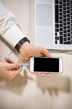 Dizüstü bilgisayarla masada oturan ve akıllı telefonunu kablolu dizüstü bilgisayara bağlayan yöneticinin yakın çekim