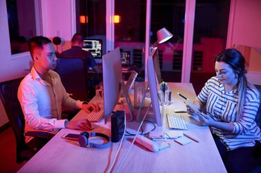 Genç Asyalı iş adamı masada oturup bilgisayarla uğraşırken, genç iş kadını karşısında oturup dijital tablet kullanırken, gece yarısına kadar ofiste çalışıyorlar.