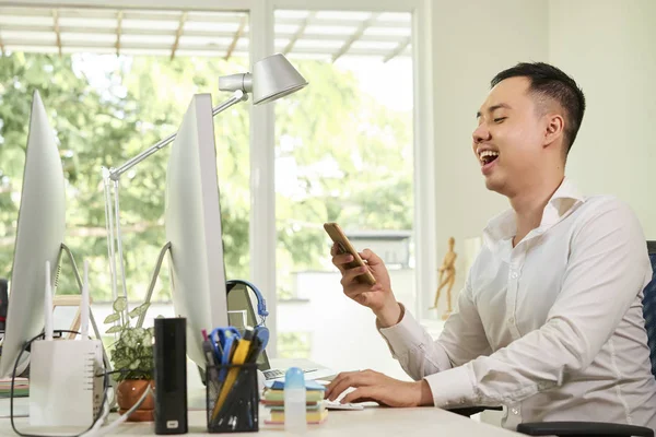 快乐的亚洲年轻人坐在桌旁 看着自己的智能手机 他在办公室工作日在网上交流 — 图库照片