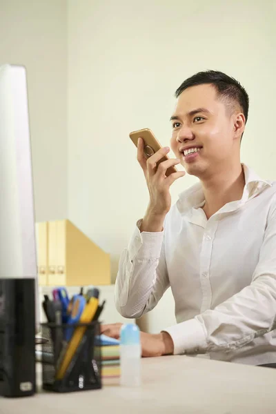 Smiling Asian businessman recording audio message on mobile phone while working with computer at his workplace at office