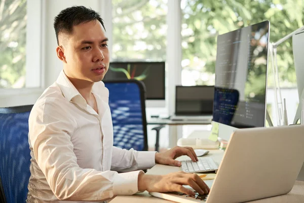严肃的亚洲经理坐在桌旁 在办公室同时配置他使用电脑和在笔记本电脑上打字的软件 — 图库照片