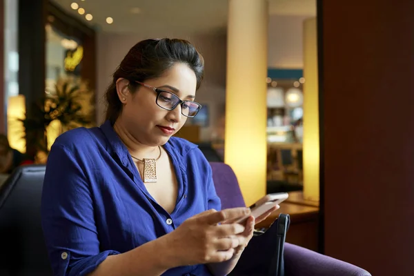 Serious young woman in eyeglasses using free internet while sitting on sofa in cafe, she reading a message on her mobile phone