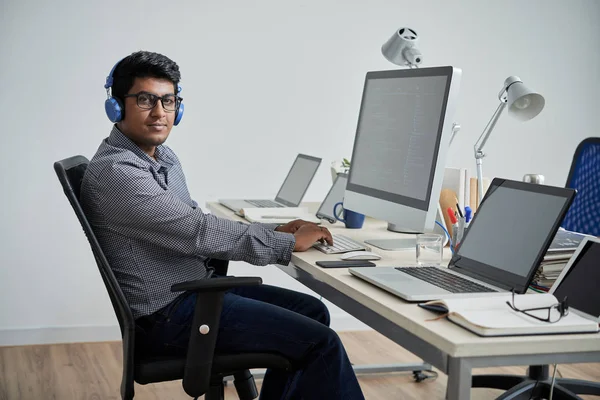 印度开发人员的肖像 戴着耳机和眼镜 在餐桌上处理电脑时看着相机 — 图库照片