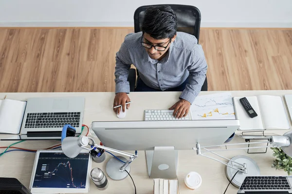 High view image of serious IT manager wearing eyeglasses sitting at the table typing on computer keyboard and looking at monitor at modern office