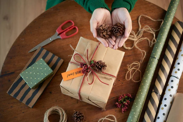 クリスマスプレゼントを準備する天然松のコーンで包まれたプレゼントをデザインする人の上から顔のないショット — ストック写真