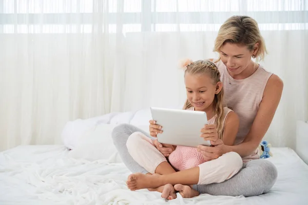 陽気な母と娘はベッドに座って デジタルタブレットで漫画を見て — ストック写真
