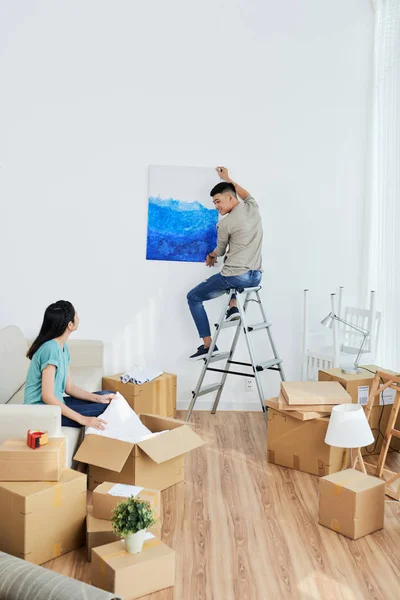 Young Asian man sitting on ladder and asking girlfriend where to hang picture while relocating into new flat