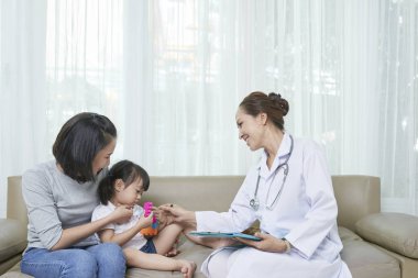 Gülümseyen çocuk doktoru kanepede oturur ken ve annesiyle doktora gelirken küçük hastayla konuşuyor.