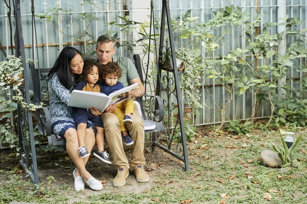 Fröhliche Mischlingsfamilie Sitzt Auf Schaukel Und Liest Zusammen Märchen — Stockfoto