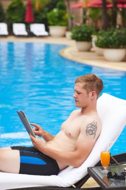 Yakışıklı genç adam yüzme havuzu tarafından dinlenme ve tablet bilgisayar üzerinde çalışıyor