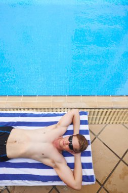 Güneş gözlüklü genç adam yüzme havuzunda çizgili havlunun üzerinde yatıyor.