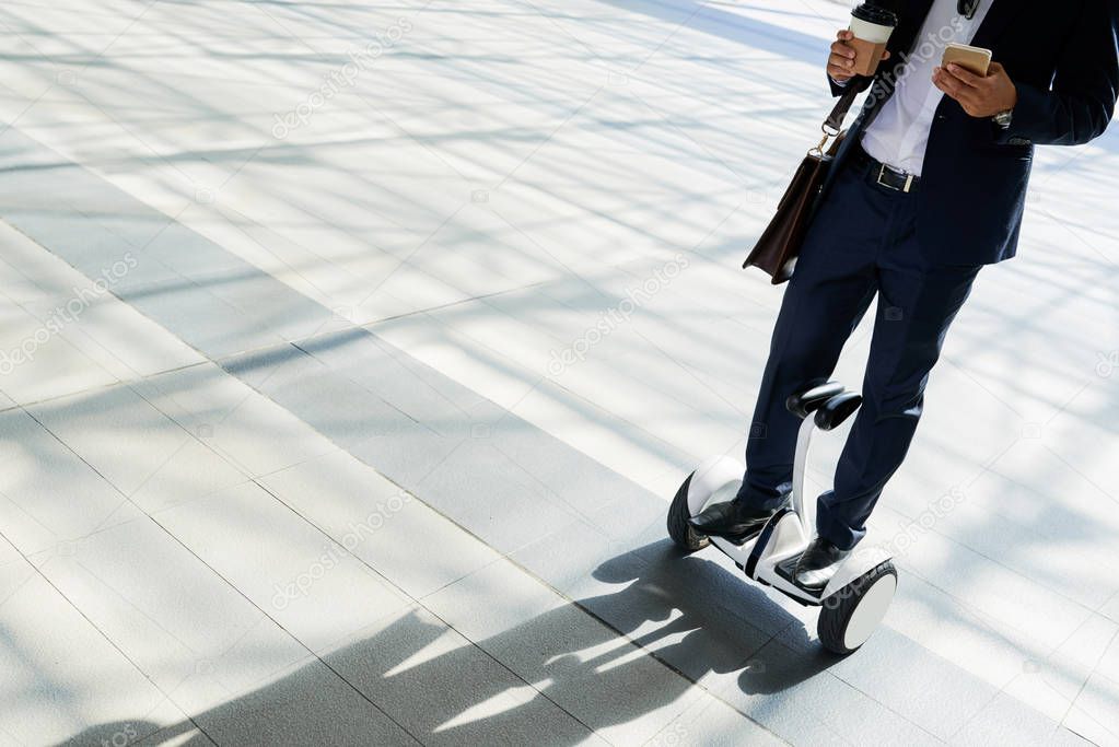 Businessman checking text messages, drinking take-out coffee and riding hoverboard