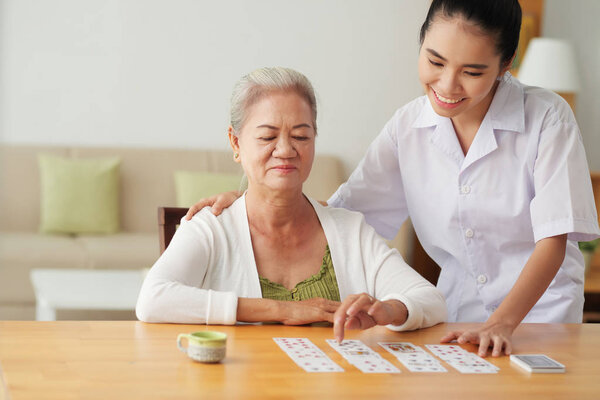 Smiling Vietnamese senior woman playing cards under supervision of skilled health worker