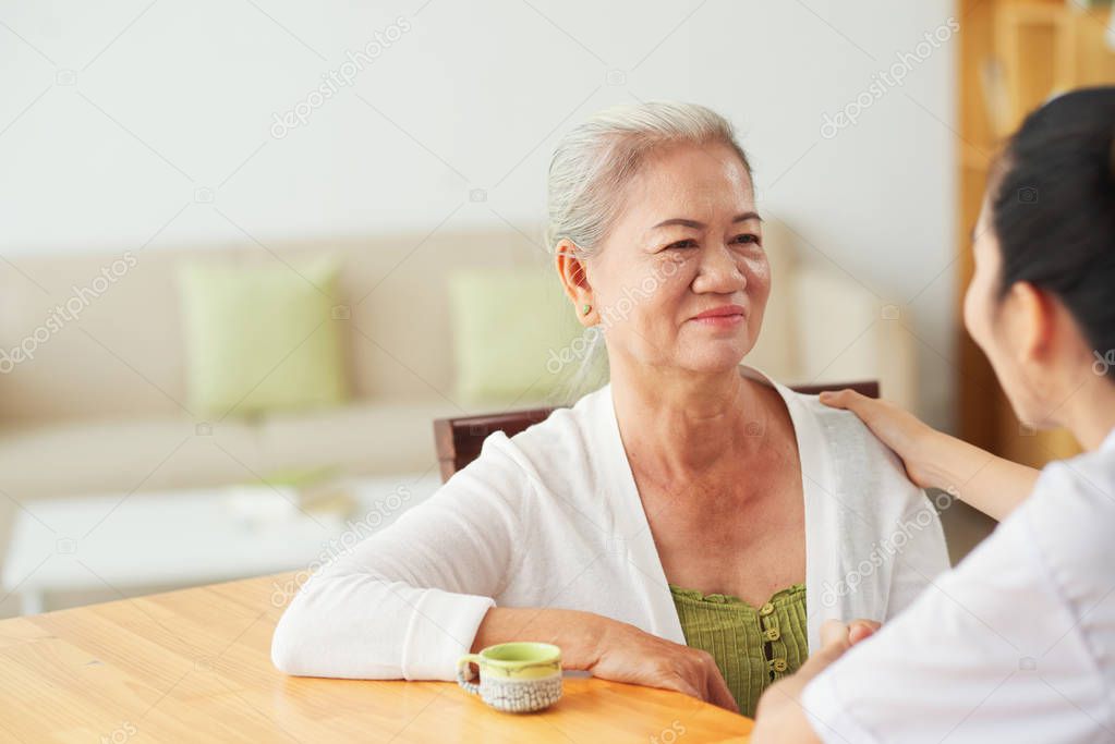 Smiling aged Asian woman drinking tea and talking to her nurse at home