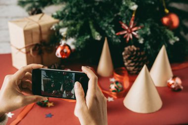 Sosyal medyada yayınlamak için ağaca karşı Noel kompozisyonu fotoğraflayan tanınmayan kadının yakın çekimi