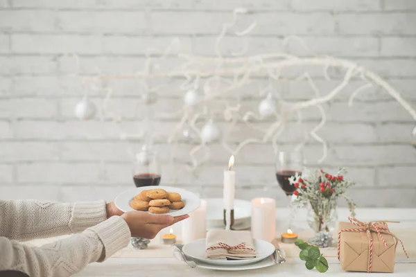 Close-up of unrecognizable woman in warm sweater putting plate with homemade cookies on table while serving it for Christmas dinner, dining table with place setting decorated candles and twigs
