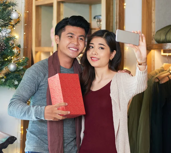 Content young Asian man and woman with Christmas presents taking selfie with phone at home