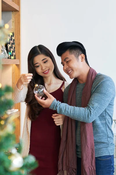 Content young Asian couple standing with glass transparent bauble at home enjoying Christmas time