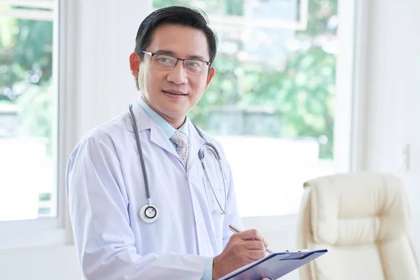 微笑的亚洲医生站在白色外套和听诊器在他的脖子上 并写一个治疗的肖像 — 图库照片