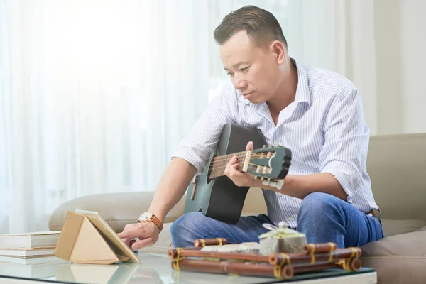 严肃的中年男子坐在坐在一个坐 并使用他的平板电脑 而在家里弹吉他 — 图库照片