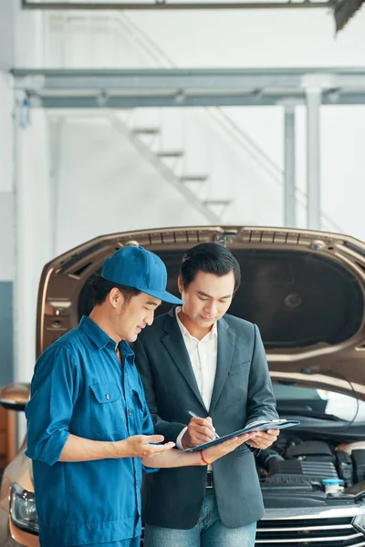 若い男は彼の近くに立って 車の問題を説明する自動車整備士と彼の車を修理する契約に署名 — ストック写真