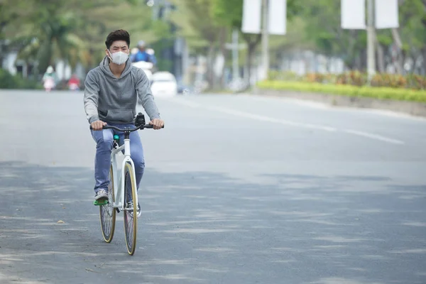 Young Vietnamese man in facemask riding on bicycle along empty road