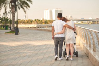 Bebek arabası ile açık havada yürüyen kucaklayan karı kocanın arka görünümü