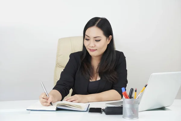 アジアの若いビジネスウーマンがラップトップの前のテーブルに座り オフィスでノートにメモを作る — ストック写真