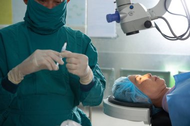Ameliyat masasında yatan olgun kadın, cerrahı ayakta durarak ve hastası için şırınga da anestezi hazırlarken lazer ekipmanı altında 