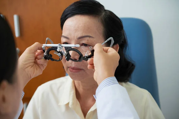 亚洲资深女子坐在视光师办公室 而医生在脸上戴视光师试验框检查视力 — 图库照片