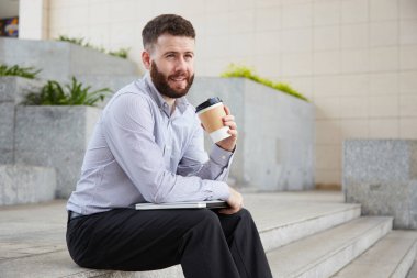 Sakallı yönetici dizlerinin üzerinde dizüstü bilgisayar ile merdivende oturan ve kahve molası açık havada sırasında tek kullanımlık fincan kahve içme