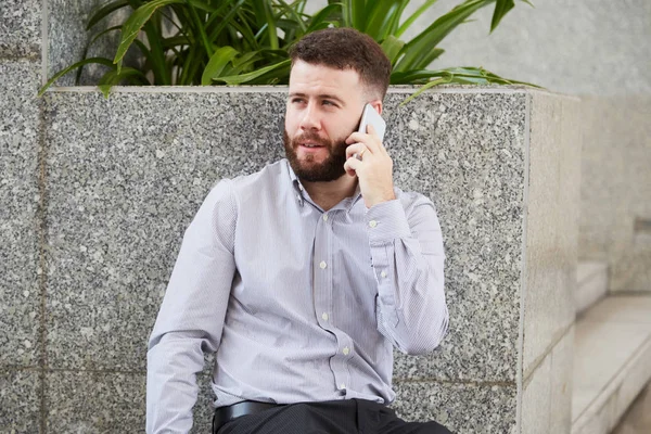 Young bearded office worker sitting outdoors in the city and talking on mobile phone