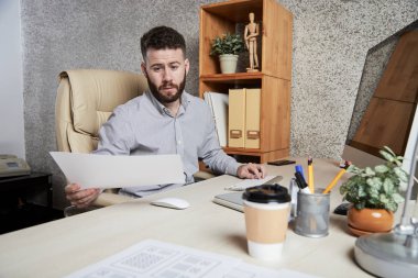 İşyerinde bilgisayar monitörünün önünde oturan ve elinde ki belgeyi ofiste okuyan ciddi sakallı işadamı