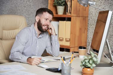 Mutlu sakallı işadamı bilgisayar monitörü bakarak ve ofiste yeni fikirler hakkında düşünme not defteri ile masada oturan 