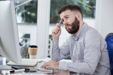 Pensive sakallı ofis çalışanı masada oturan ve bilgisayar üzerinde çalışırken yeni iş projesi hakkında düşünme