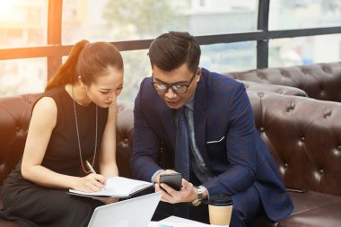 Kanepede oturan iki iş ortağı birlikte akıllı telefona bakıyor ve ofiste ki iş sırasında not defterine notlar alıyor