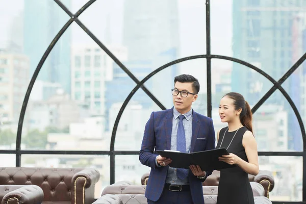 アジアのビジネスカップルは ドキュメントと黒いフォルダとフォーマルな服を着て立って 大きな窓を持つオフィスで何かを見て — ストック写真